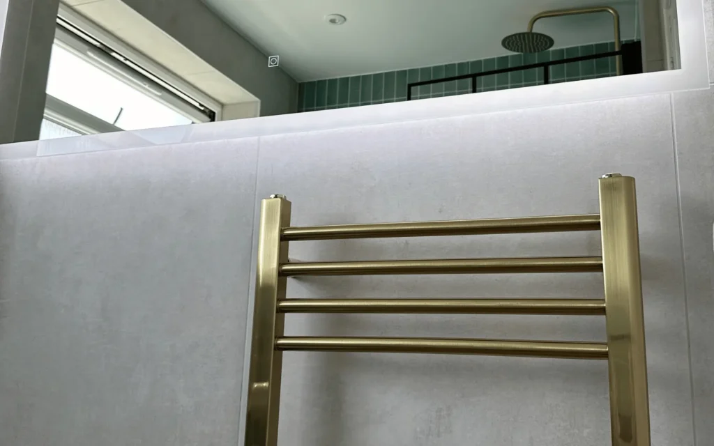 modern brushed brass radiator installation with backlit mirror and grey concrete effect bathroom tiling.
