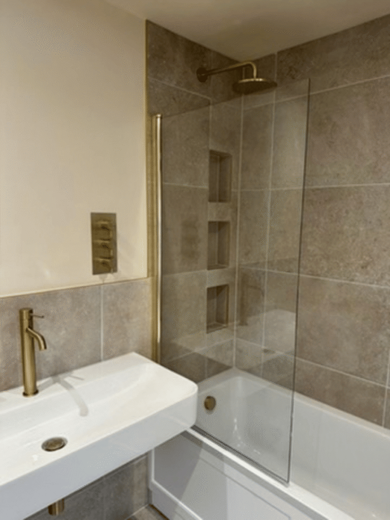 Complete bathroom fitting in Taunton featuring a shower over bath, brushed brass fittings, glass shower screen and grey stone-effect tiles by Plumb Inc.