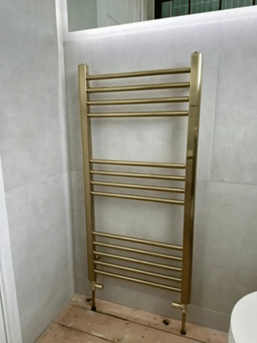 Brushed brass heated towel rail radiator installed by Plumb Inc in Taunton, featuring a ladder-style design against contemporary grey bathroom wall tiles.