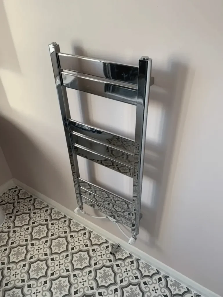 Chrome heated towel rail radiator installed by Plumb Inc in Taunton, featuring a polished chrome finish and patterned bathroom floor tiles.